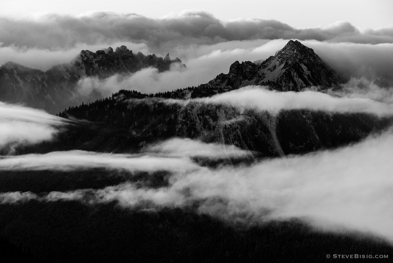 A black and white photograph of mountain ranges surrounding Mount Rainier peaking through the clouds on an Autumn day. Mount Rainier National Park, Washington.