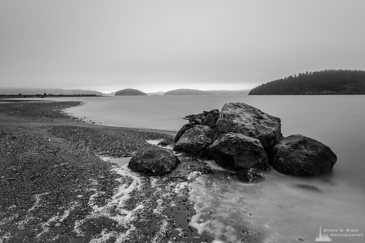 A black and white, long exposure landscape photograph of a group of rocks on the coastline at Ala Spit on Whidbey Island, Washington.