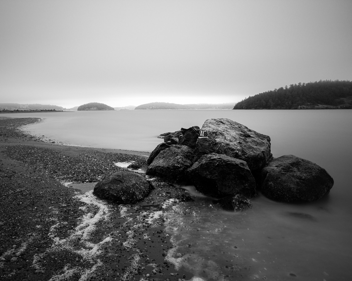 A black and white, long exposure landscape photograph of a group of rocks on the coastline at Ala Spit on Whidbey Island, Washington.