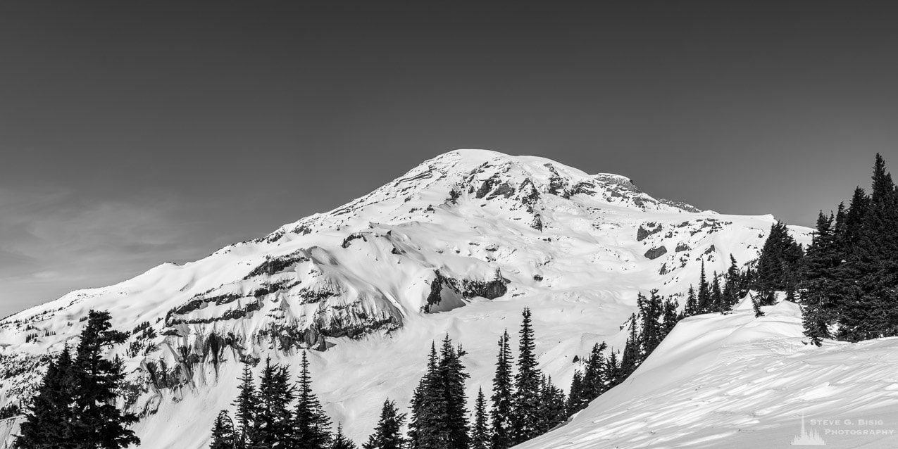A black and white panoramic landscape photograph of Mount Rainier as viewed from the snow covered meadows near the Paradise Visiitors Center at Mount Rainier National Park, Washington.
