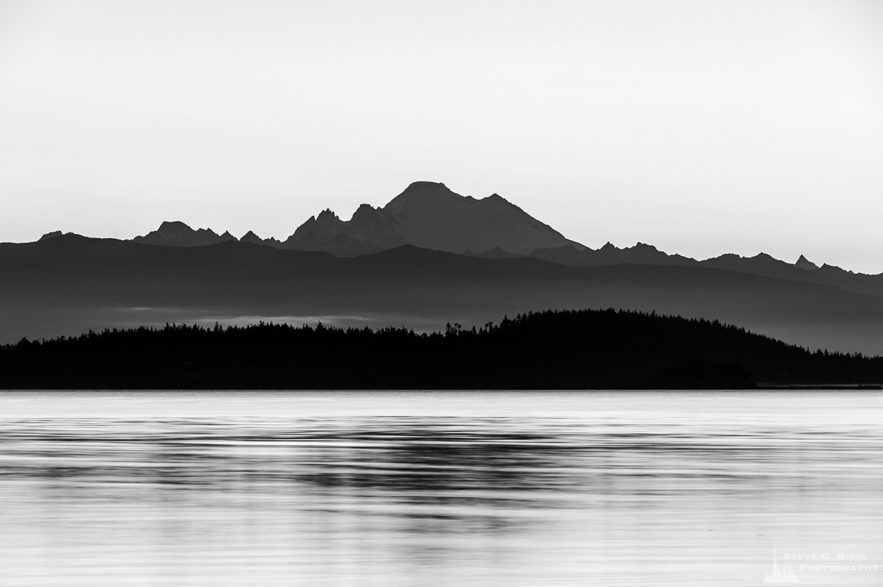 A black and white landscape photograph of Mount Baker in the early morning Summer light over Skagit Bay as seen from Borgman Road on Whidbey Island near Oak Harbor, Washington.
