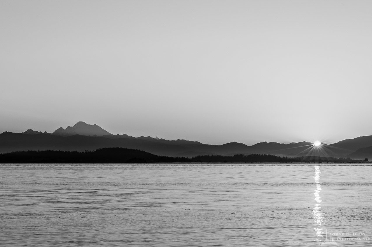 A black and white landscape photograph of the Summer sunrise over the Cascade Mountains and Skagit Bay as seen from Borgman Road on Whidbey Island near Oak Harbor, Washington.