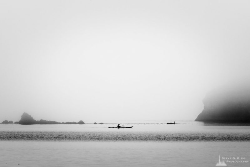A black and white landscape photograph a lone kayaker on Bowman Bay on a foggy Summer morning at Deception Pass State Park, Washington.
