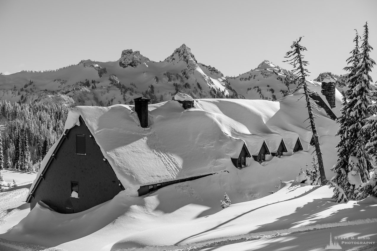 A black and white photograph of the historic Paradise Inn under a heavy blanket of snow as captured on a sunny winter day in the Paradise area of Mount Rainier National Park, Washington.