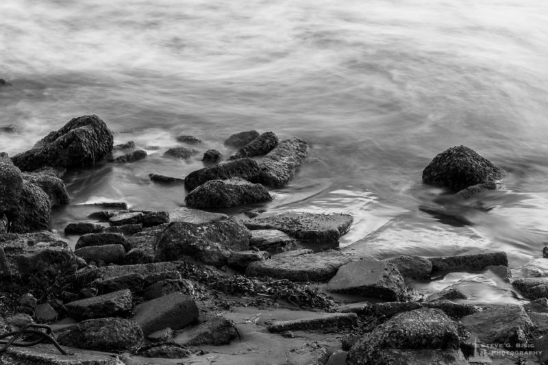 A long exposure black and white landscape photograph of the waves churning along the rocks at North Cove, Washington.