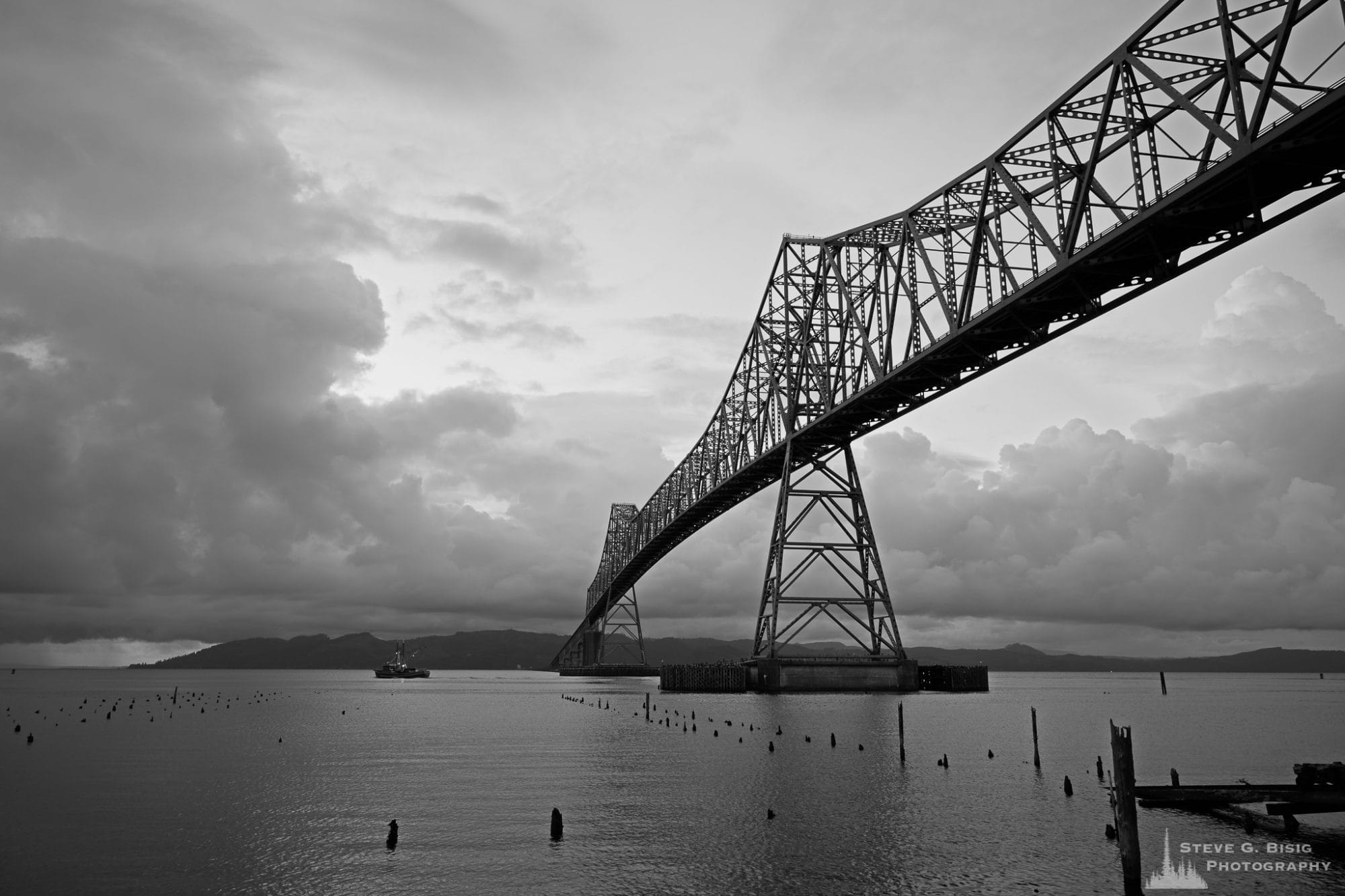 A black and white landscape photograph of the Astoria-Megler Bridge over the Columbia River on a dark, cloudy summer day as viewed from Astoria, Oregon.