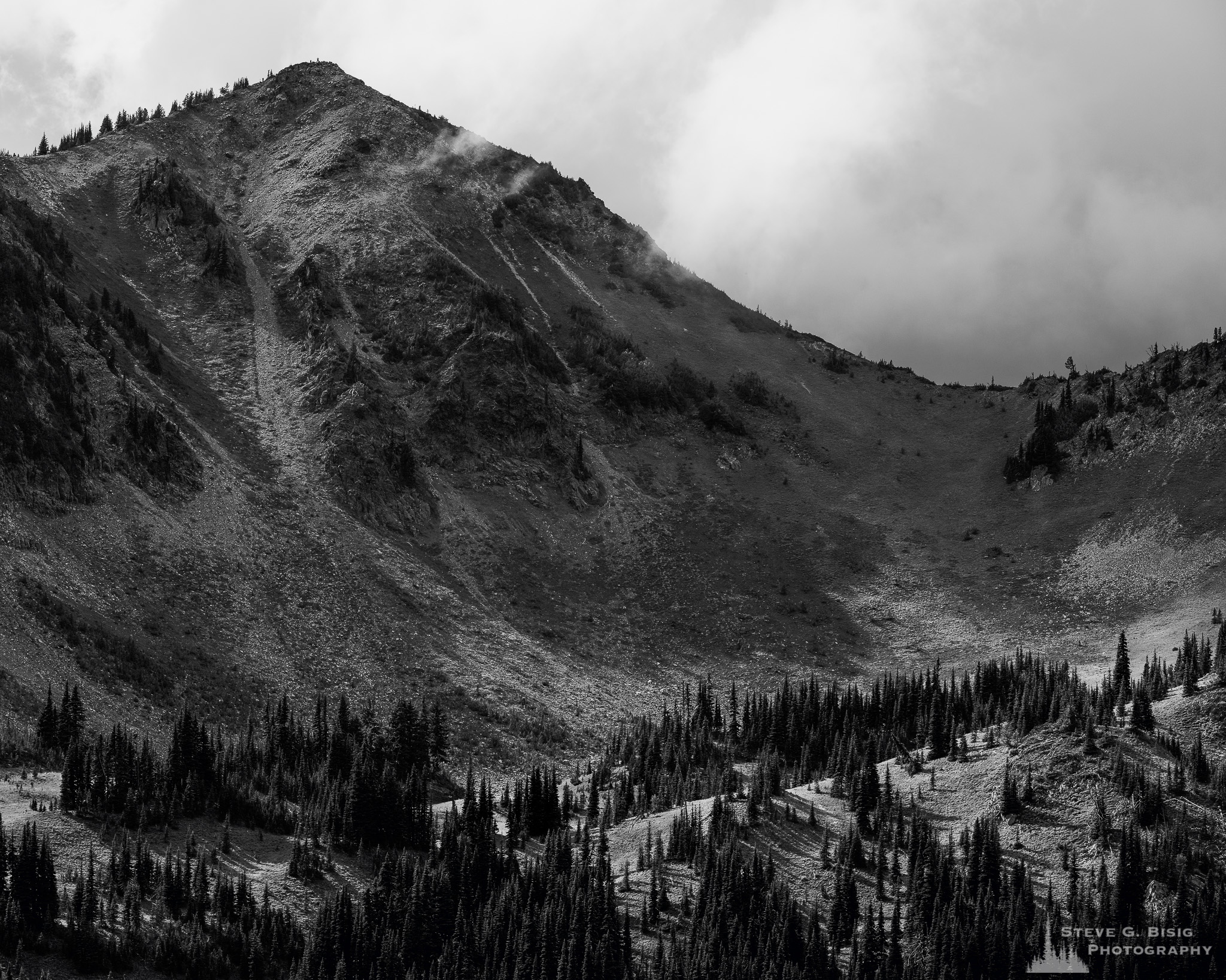 A black and white landscape photograph of Dege Peak on a cloudy late summer day with the sun highlighting the meadows below at Mount Rainier National Park, Washington.