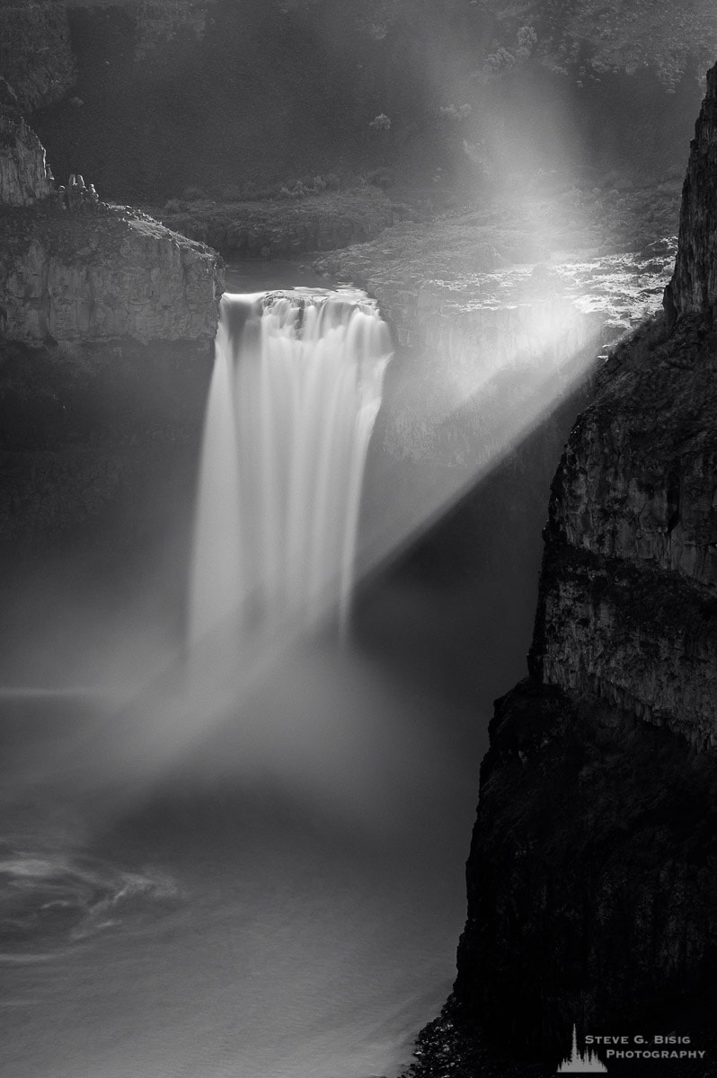 A black and white landscape photograph of Palouse Falls with light rays from the backlit sunlight.