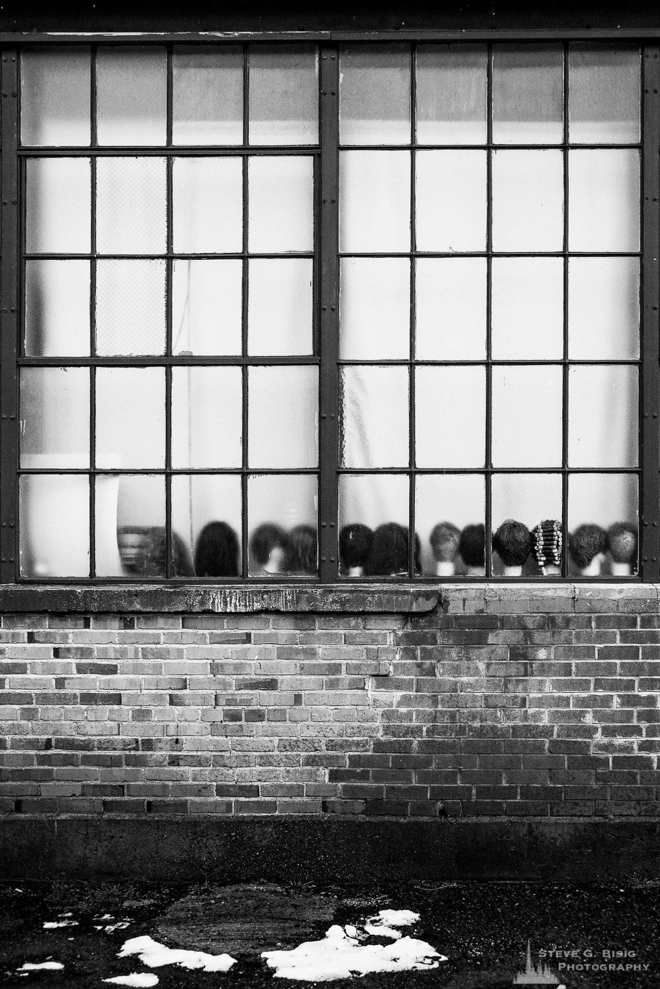 A black and white urban photograph of the window of an old brick building housing a beauty school in Ellensburg, Washington with wig heads on the ledge.