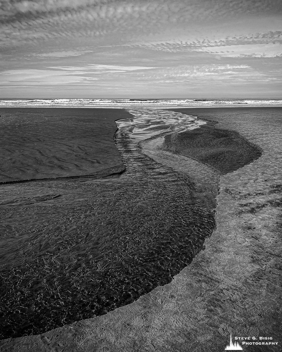 A black and white mobile photograph of a creek flowing across the beach out into the Pacific Ocean at Cannon Beach, Oregon.