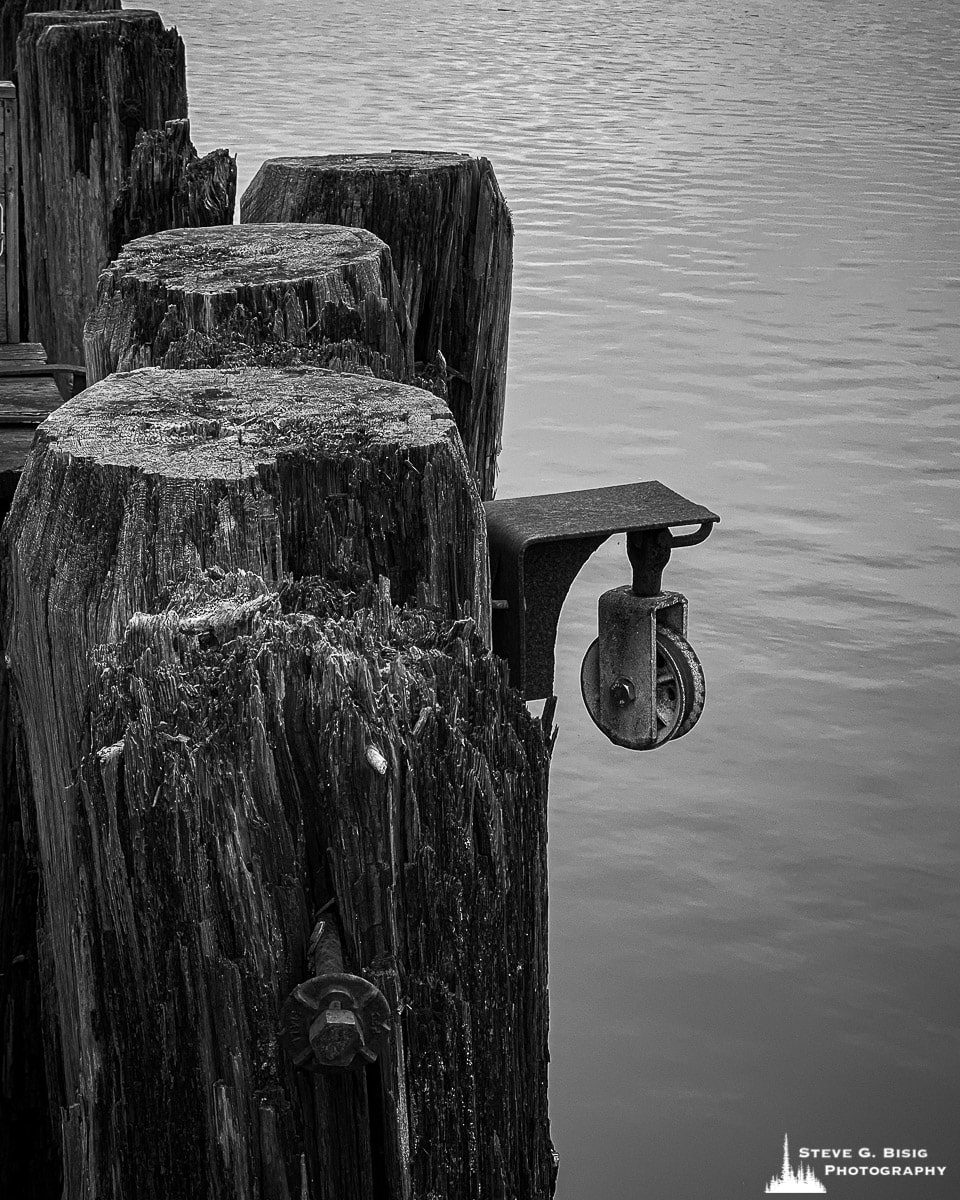 A black and white mobile photograph of an old pully on a dock along the historic waterfront at Astoria, Oregon.