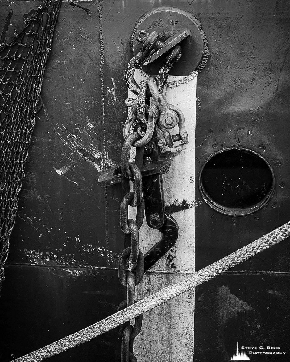 A black and white mobile photograph of a porthole, chain, net, and rope on the side of Lightship Columbia moored along the historic waterfront in Astoria, Oregon.