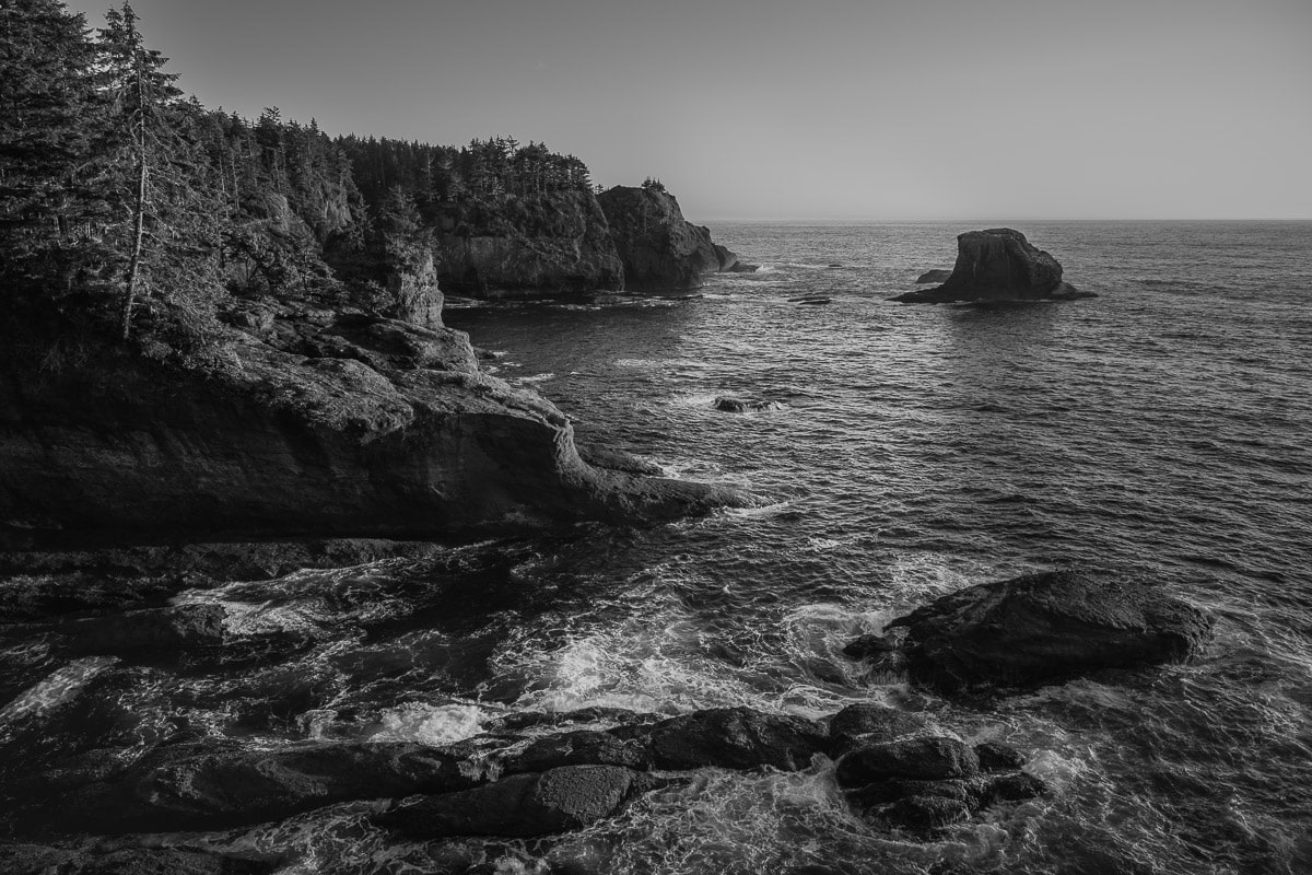 A black and white landscape photograph of the summer evening lighting shining on the coastline over the Pacific Ocean at Cape Flattery, Washington.