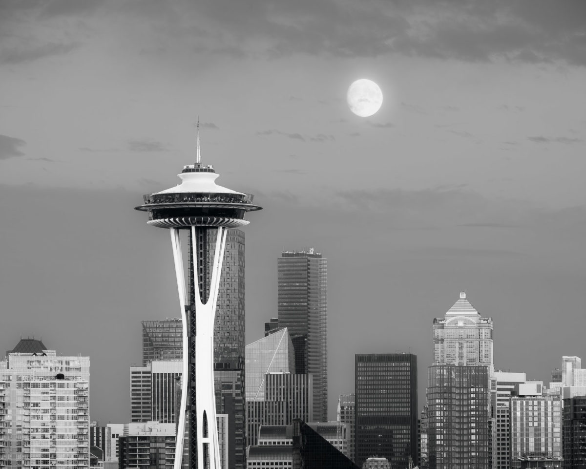 A black and white urban photograph of the full moon over the Space Needle and downtown Seattle, Washington.