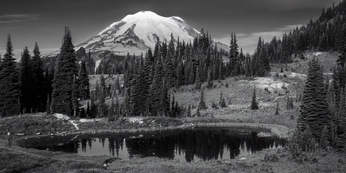A black and white panoramic landscape photograph of Mt. Rainier over looking an alpine pond as viewed along the Naches Peak Loop trail near Chinook Pass, Washington.