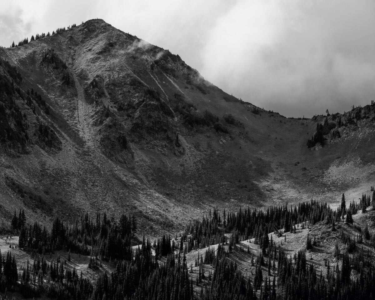A black and white landscape photograph of Dege Peak on a cloudy late summer day with the sun highlighting the meadows below at Mount Rainier National Park, Washington.