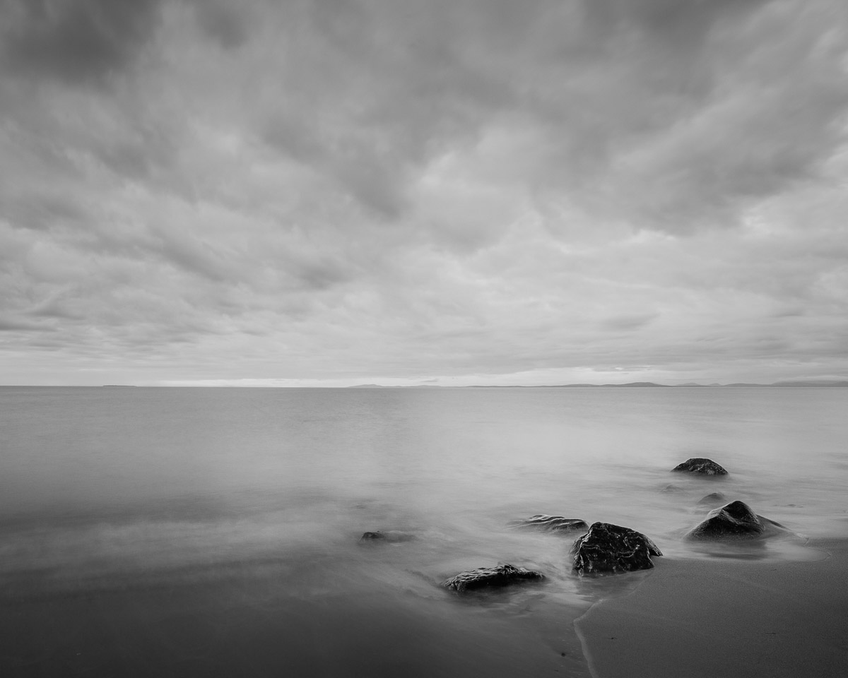 Under the dramatic dance of early autumn clouds, the serene waters along West Beach on Whidbey Island, Washington, mirror the skies’ ethereal beauty. The monochromatic tones capture a moment of stillness where rocks silently witness nature’s artistry. It’s a scene where the elements of earth, water, and sky converge in harmonious contrast.