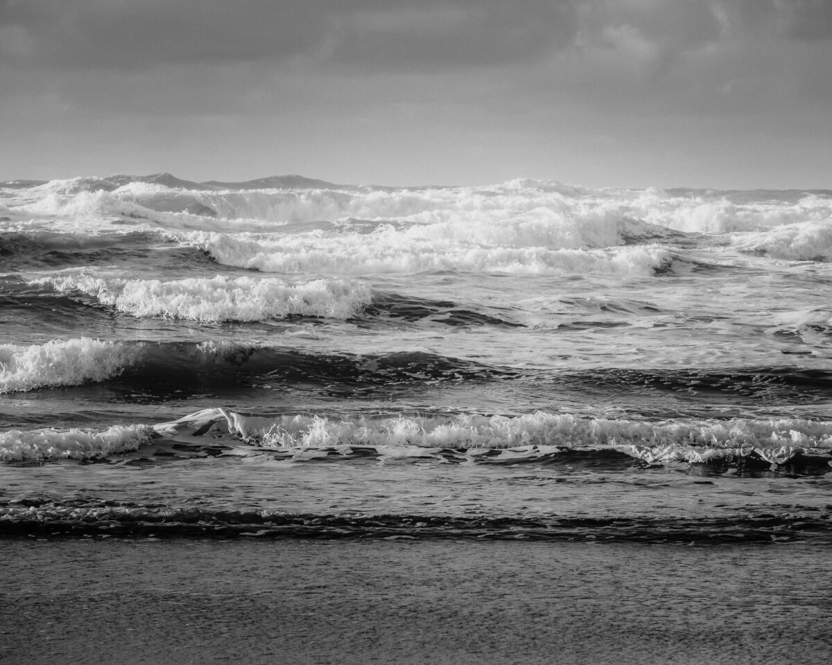 A black and white photo of the ocean's waves breaking onto the shore. The photo conveys the soothing and calming sound of the ocean's song. The waves are rhythmic and harmonious, with the horizon creating a sense of infinity.