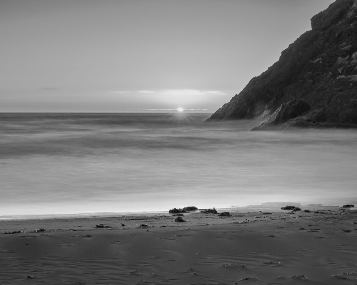 A black and white landscape photograph of the summer sunset over the Pacific Ocean at Deadmans Hollow, Cape Disappointment State Park near Ilwaco, Washington.