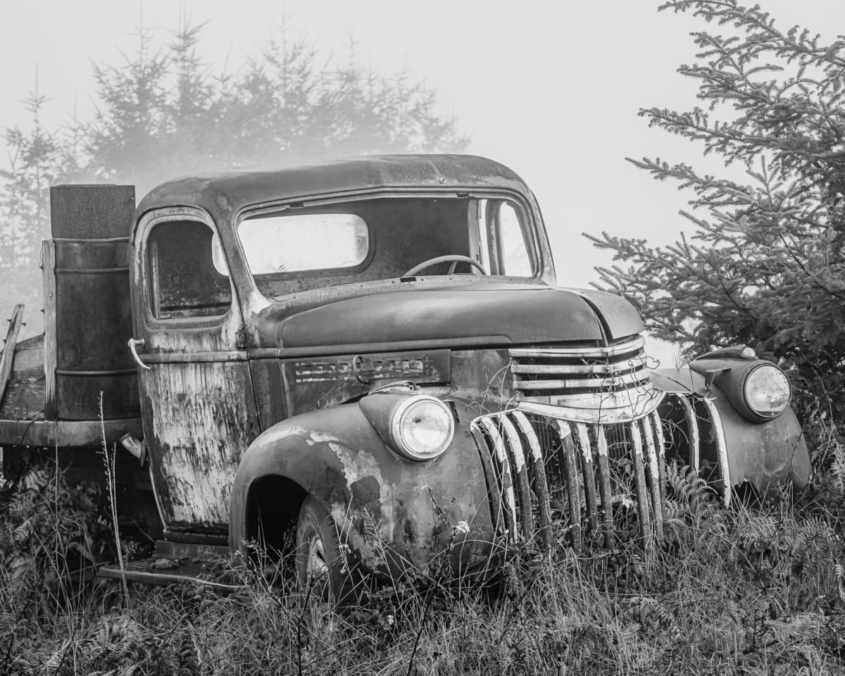 This black and white photograph captures a rusty Chevrolet flatbed farm truck, retired and succumbing to the elements. The old truck, a silent witness to bygone days, is surrounded by overgrown grass and backed by misty trees, evoking a sense of nostalgia. The rust and decay on the truck paint a poignant picture of nature reclaiming its space.