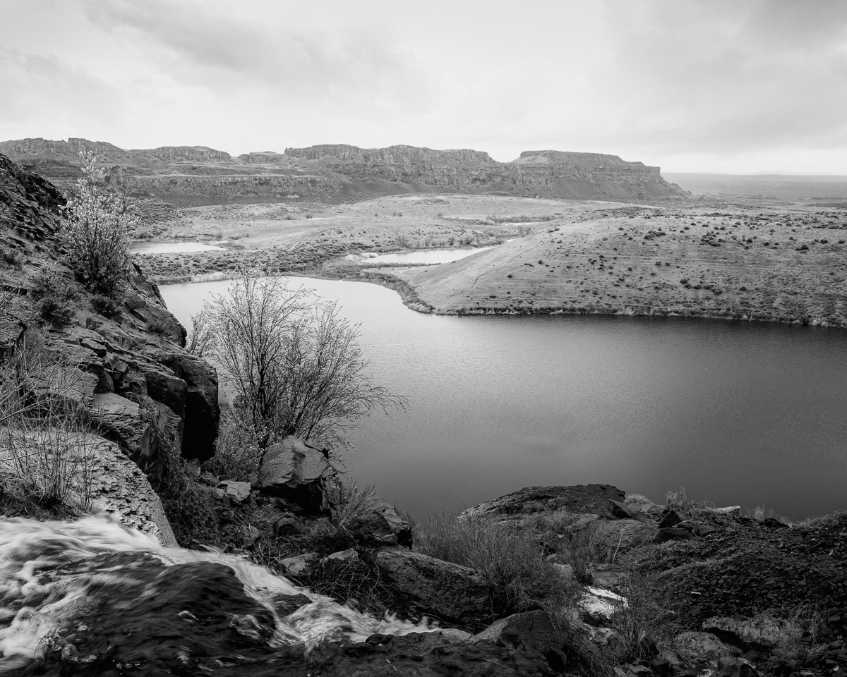 A black and white landscape photograph looking over the edge of a waterfalls as it plunges into the Potholes Coulee above the Ancient Lakes area in the Quincy Lakes Unit of the Columbia Basin Wildlife Area in Grant County, Washington.