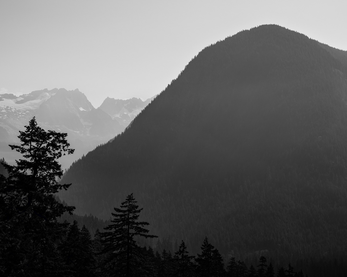 In the serene Cascade River Valley of Washington State, the imposing silhouette of a forested mountain dominates the scene, with distant peaks barely visible through the haze of wildfire smoke. The play of light and shadow, filtered by the smoky air, evokes a sense of tranquility as the rugged landscape rises from the mist. This timeless moment captures the stillness of the North Cascades, softened by nature's unpredictability, and showcases the enduring beauty of our natural world.