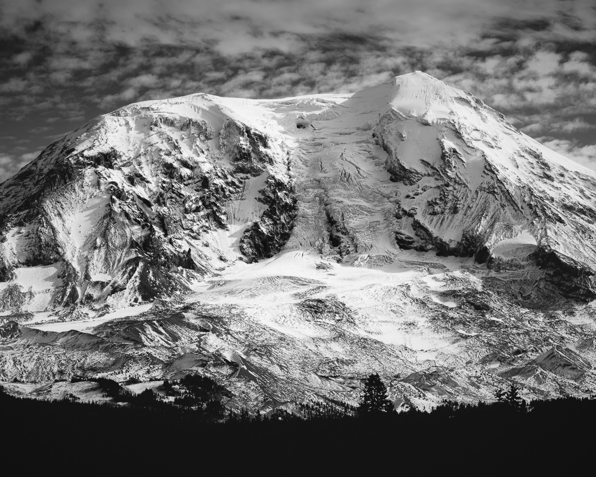 A black and white landscape photograph capturing the imposing form of Mt. Adams, cloaked in a fresh Autumn snow. The interplay of light and shadow highlights the mountain's grandeur and the serene beauty of the surrounding landscape.