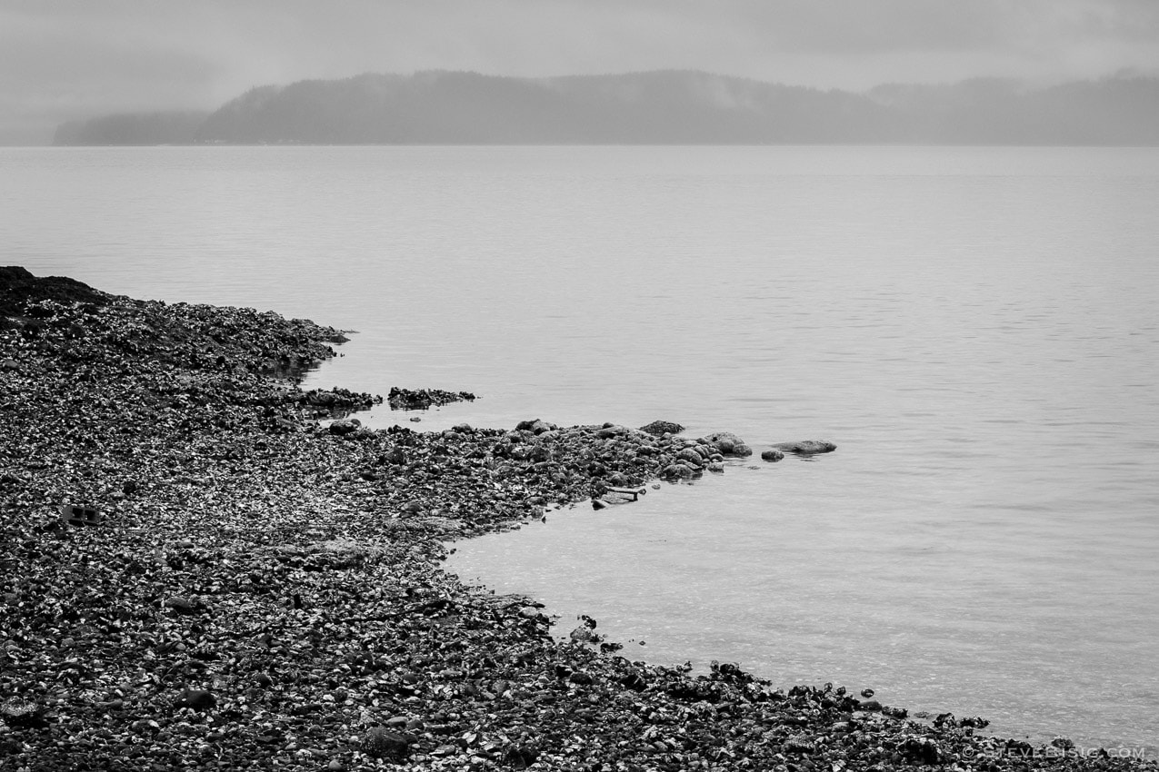 A black and white photograph of the rocky beach along Hood Canal at the Triton Cove State Park near Brinnon, Washington. The Kitsap Peninsula in the distance.